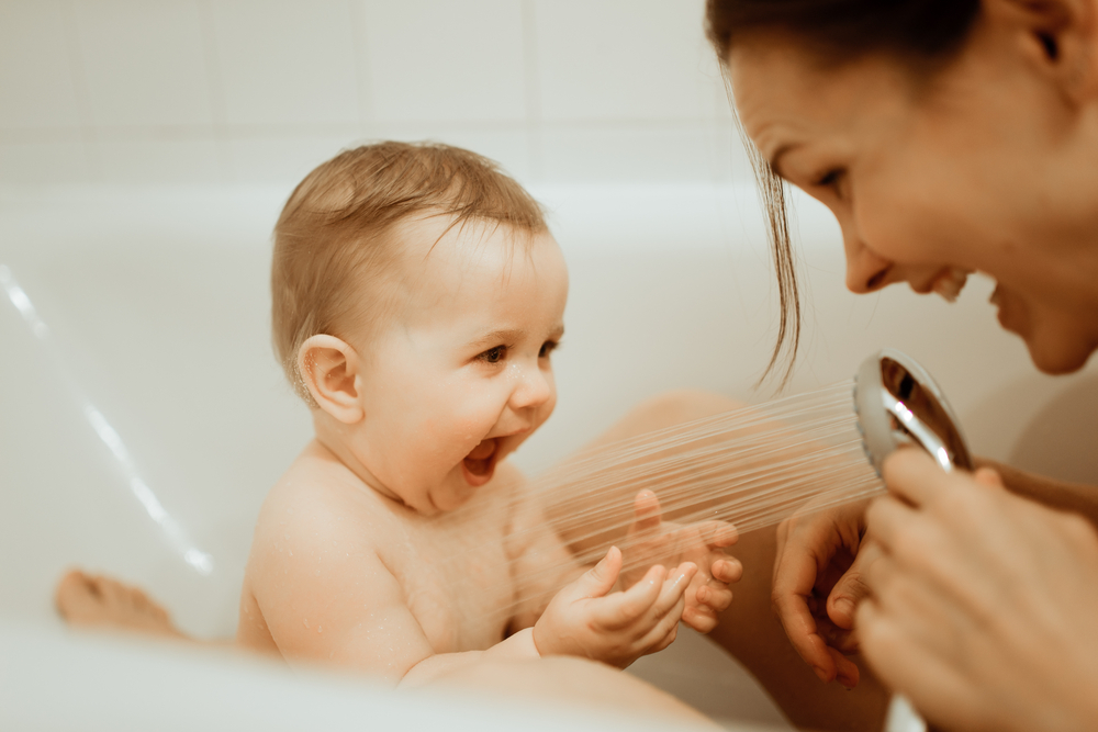 Ein Baby sitzt in der Badewanne und wird mit dem Duschkopf von seiner Mama abgeduscht.