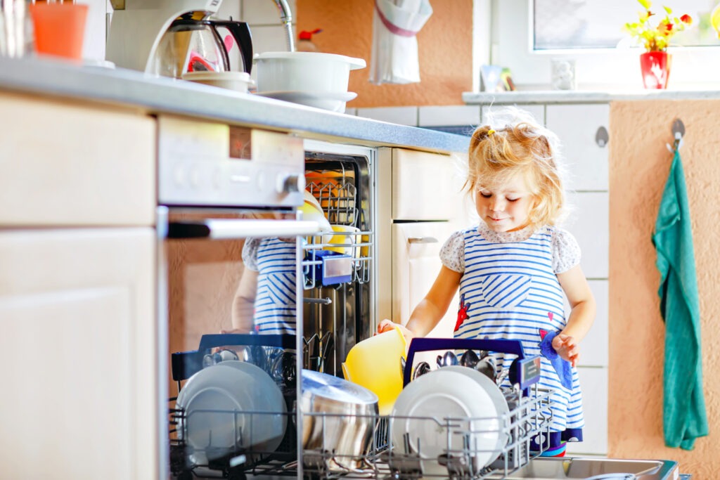 Kleine, niedliche Kleinkind-Mädchen hilft dabei, den Geschirrspüler auszuräumen – ein lustiger Moment, der zeigt, wie Kinder im Haushalt helfen können.

