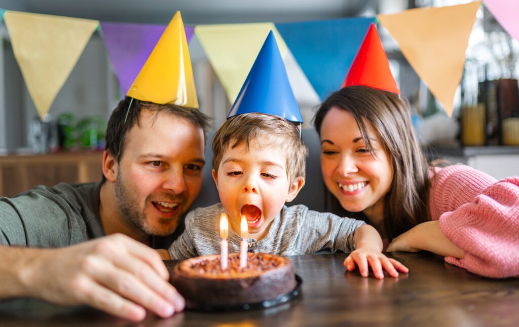 Eine Familie feiert den zweiten Geburtstag ihres kleinen Sohnes. Die Eltern stehen lächelnd neben dem Jungen, der vor einem bunt dekorierten Geburtstagskuchen sitzt.Sinnvolle und besondere Geschenkideen für den 2. Geburtstag