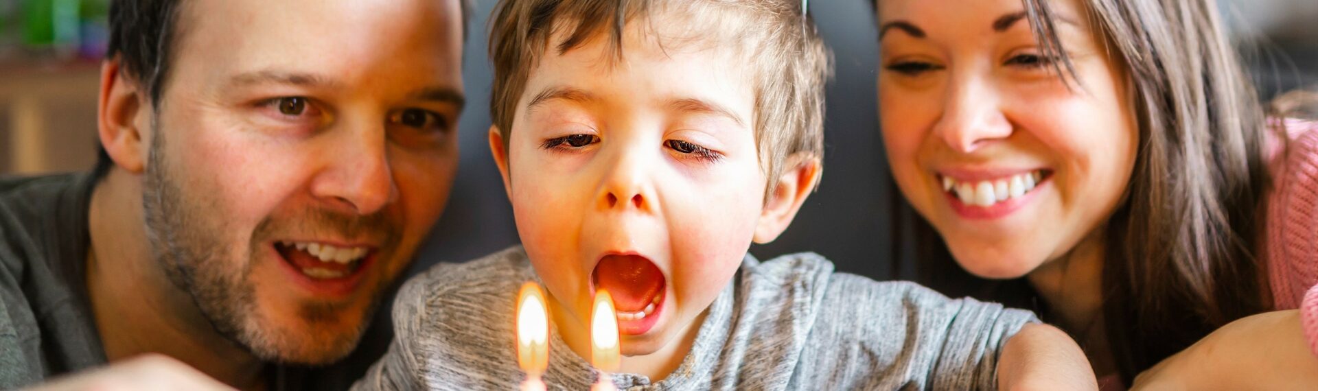 Eine Familie feiert den zweiten Geburtstag ihres kleinen Sohnes. Die Eltern stehen lächelnd neben dem Jungen, der vor einem bunt dekorierten Geburtstagskuchen sitzt.