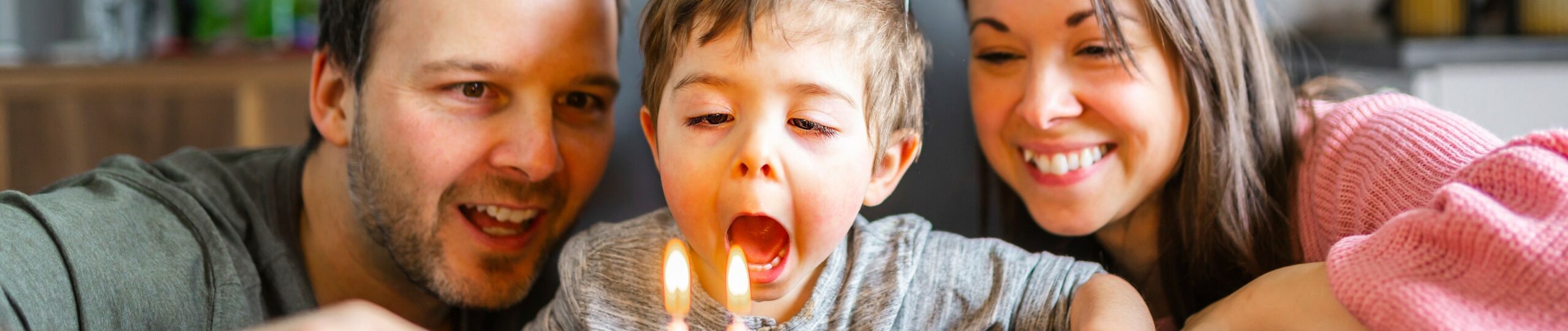 Eine Familie feiert den zweiten Geburtstag ihres kleinen Sohnes. Die Eltern stehen lächelnd neben dem Jungen, der vor einem bunt dekorierten Geburtstagskuchen sitzt.