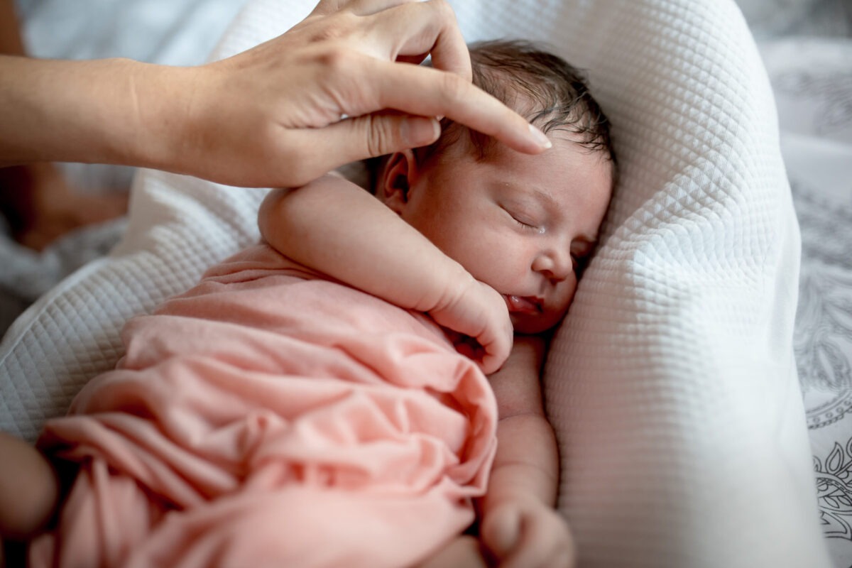 Ein Neugeborenes liegt entspannt und schlafend in einem Babynestchen und wird liebevoll im Gesicht gestreichelt.