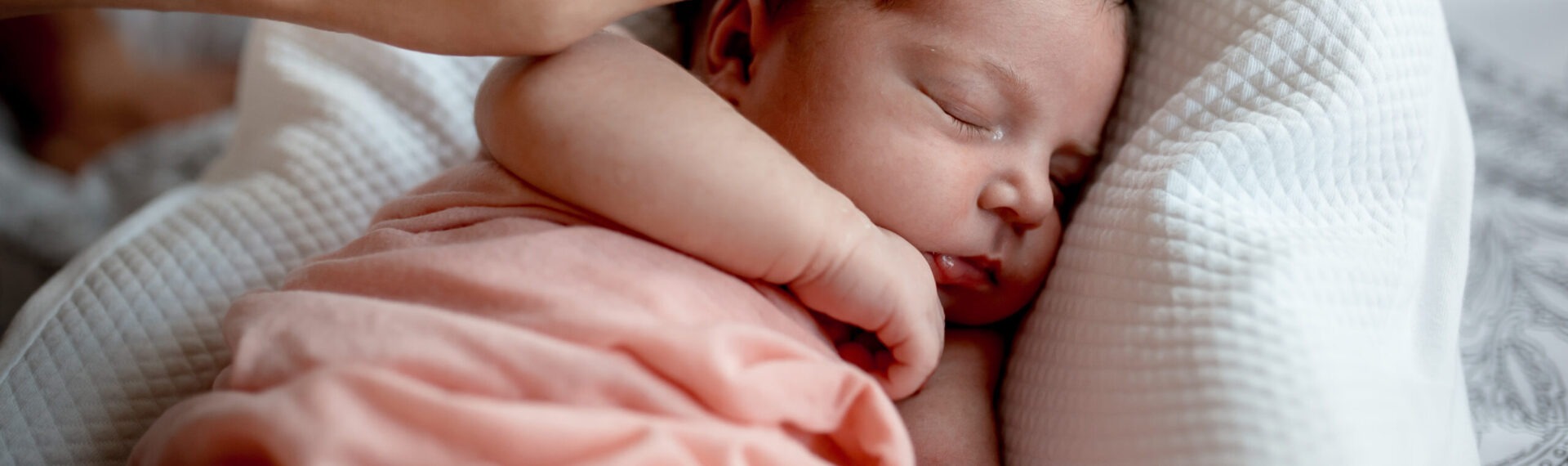 Ein Neugeborenes liegt entspannt und schlafend in einem Babynestchen und wird liebevoll im Gesicht gestreichelt.