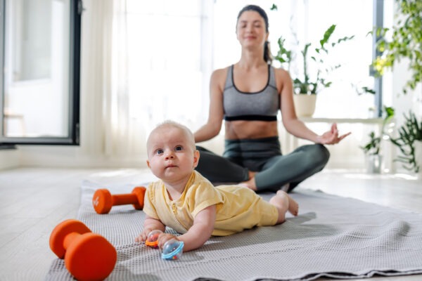 Junge Mutter meditiert in Sportkleidung mit Baby vor sich.