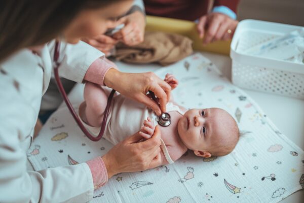 Ein Baby liegt auf einen Mull-Tuch auf einem Tisch und bekommt von einer Ärztin mit einem Stethoskop die Brust abgehört.