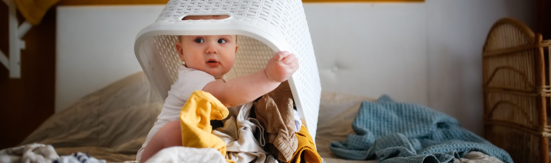 Baby sitzt unter einem ausgeleerten Wäschekorb auf dem Bett und schaut neugierig über die Schulter.
