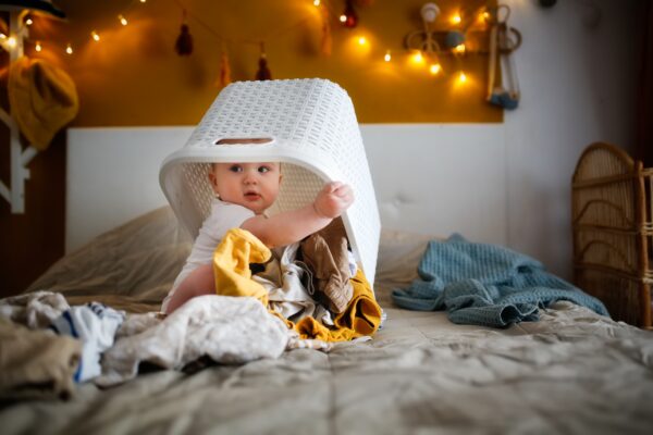 Baby sitzt unter einem ausgeleerten Wäschekorb auf dem Bett und schaut neugierig über die Schulter.