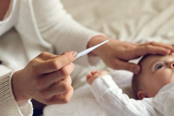 Frau misst die Temperatur eines Babys mit einem Thermometer, während sie die Hand auf dessen Stirn legt.