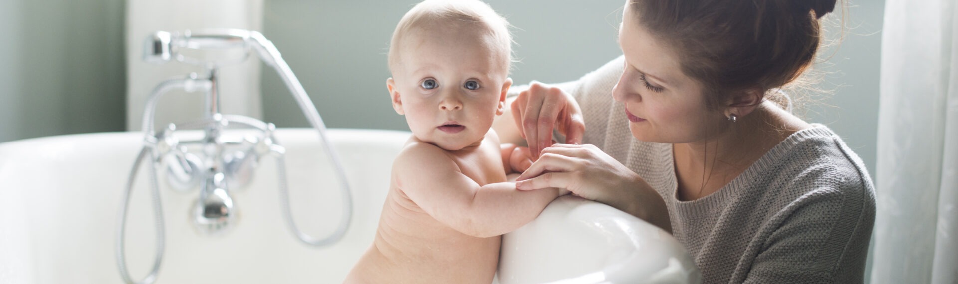 Baby sitzt in einer Badewanne, während eine Frau am Rand liebevoll seine Hand hält.