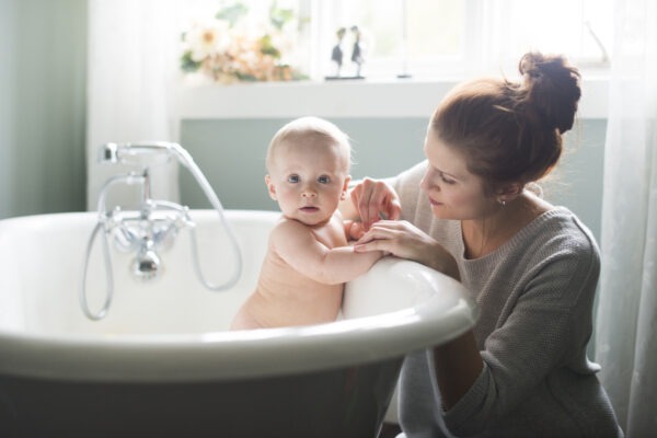 Baby sitzt in einer Badewanne, während eine Frau am Rand liebevoll seine Hand hält.