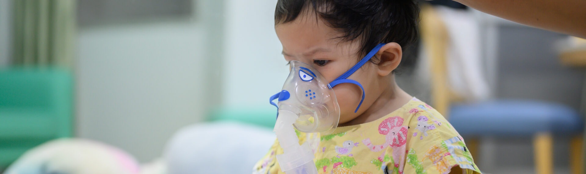 Kleines Kind mit Sauerstoffmaske sitzt auf einem Bett, während eine Hand sanft den Kopf berührt.