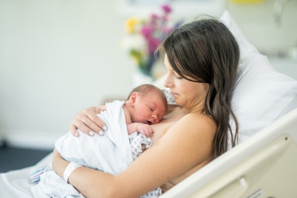 Eine Frau liegt in einem Krankenhausbett und hält ihr neugeborenes Baby liebevoll in den Armen, während das Baby auf ihrer Brust schläft.