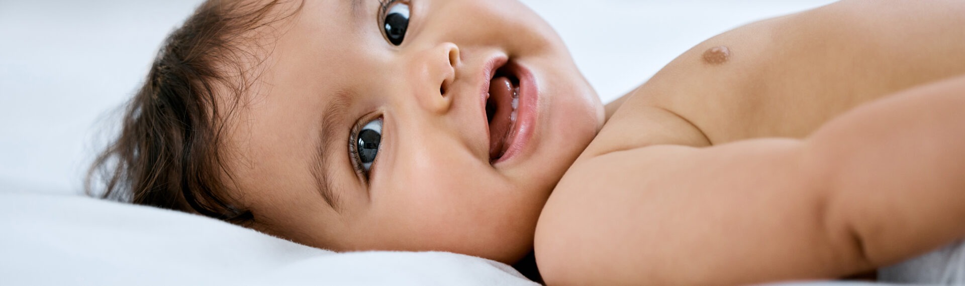 Baby liegt auf dem Bett und lächelt in die Kamera, zwei kleine Zähne blitzen auf.