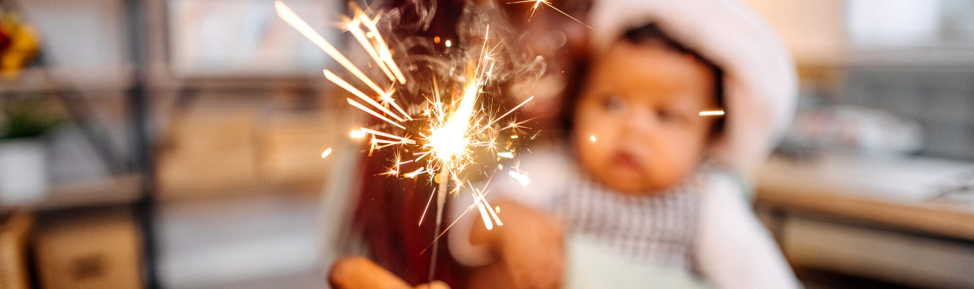 Eine Mutter schaut mit ihrem Baby eine Wunderkerze an. Mutter und Baby sind in der Unschärfe zu sehen.
