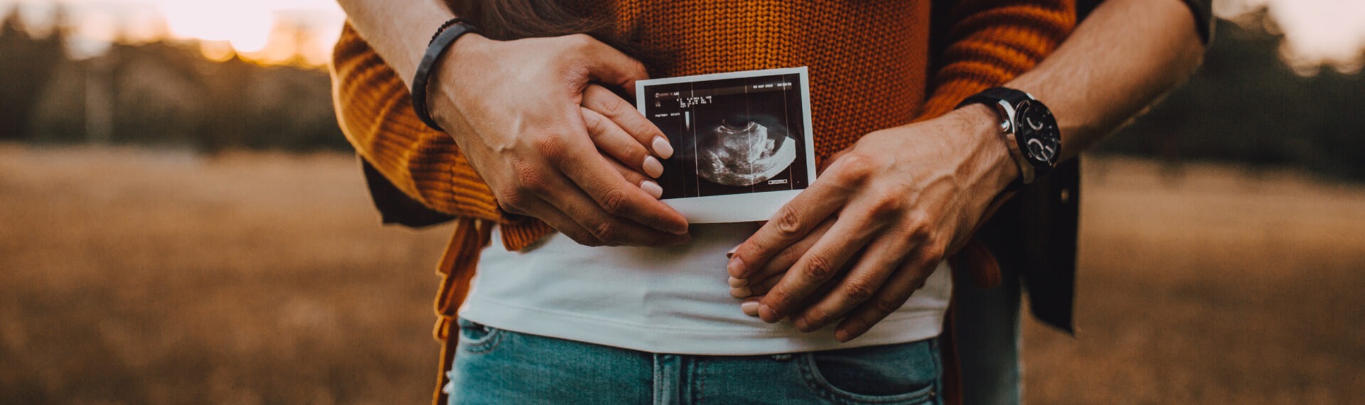 Ein Paar hält gemeinsam ein Ultraschallbild vor dem Bauch der werdenden Mutter, im Hintergrund ist eine ländliche Szene bei Sonnenuntergang zu sehen.