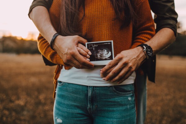 Ein Paar hält gemeinsam ein Ultraschallbild vor dem Bauch der werdenden Mutter, im Hintergrund ist eine ländliche Szene bei Sonnenuntergang zu sehen.